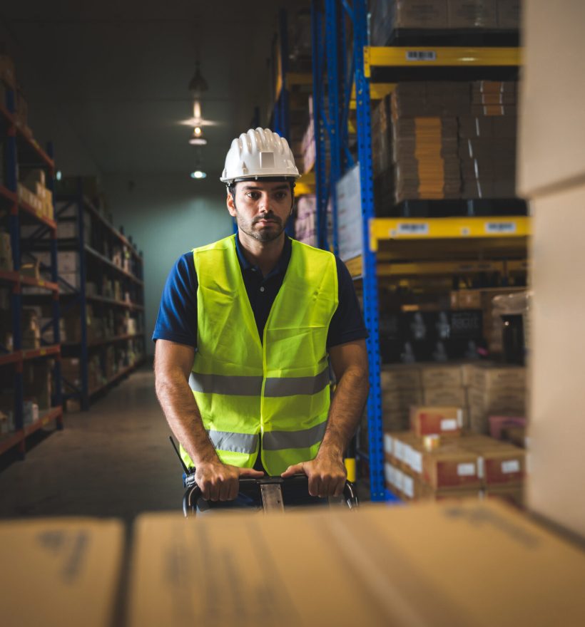 Handsome young male employee concentrating and focusing on work wearing safety helmet and uniform in warehouse while standing between racks in aisle